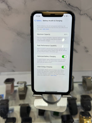 A black smartphone displaying battery health and charging settings, held upright in a silver display stand on a light marble surface with blurred watches in the background.