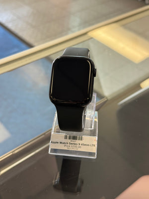 A black Apple Watch Series 9 with a black band, displayed on a clear plastic hanger with a product tag attached, resting on a reflective dark glass surface.