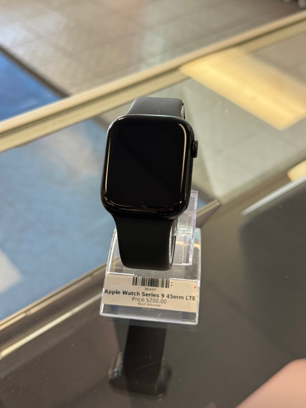 A black Apple Watch Series 9 with a black band, displayed on a clear plastic hanger with a product tag attached, resting on a reflective dark glass surface.
