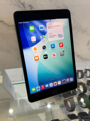 A black Apple iPad tablet standing upright on a display stand, showing the home screen with various app icons. The tablet is photographed in a retail or showroom setting with a marble-patterned background and other devices partially visible in the foreground.