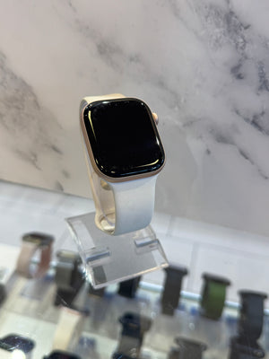 A smartwatch with a black rectangular screen and a light beige or off-white band, displayed on a clear stand in front of a marble-patterned background.
