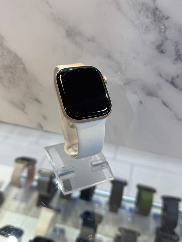A smartwatch with a black rectangular screen and a light beige or off-white band, displayed on a clear stand in front of a marble-patterned background.