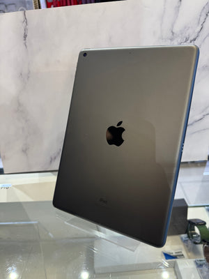 An Apple iPad shown from the back, featuring a dark gray metallic finish with the iconic black Apple logo in the center. The device is photographed standing upright on a glass display stand against a white marble-patterned background.