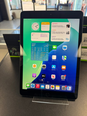 A black Apple iPad tablet standing upright on a clear acrylic display stand on a dark countertop, with the home screen visible. The background shows a blurred retail or office environment with boxes and equipment.