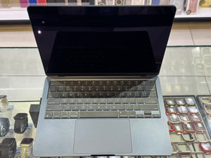 A silver laptop with a black screen and gray keyboard, displayed on a glass counter in a retail setting with watches and accessories visible in the background.