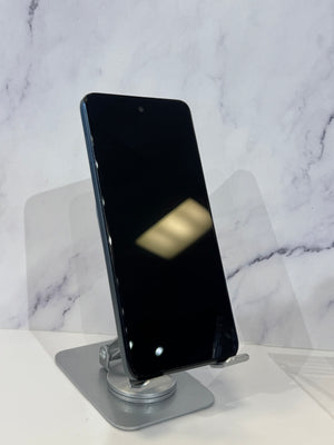 A black smartphone with a front-facing camera cutout stands upright on a silver metal display stand against a white marble-patterned background.