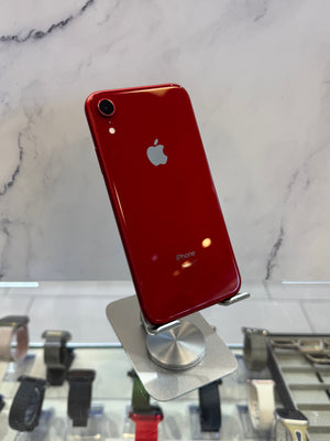 A red Apple iPhone XR standing upright on a silver display stand, showing the back of the phone with the Apple logo and camera lens visible, set against a marble-patterned background.