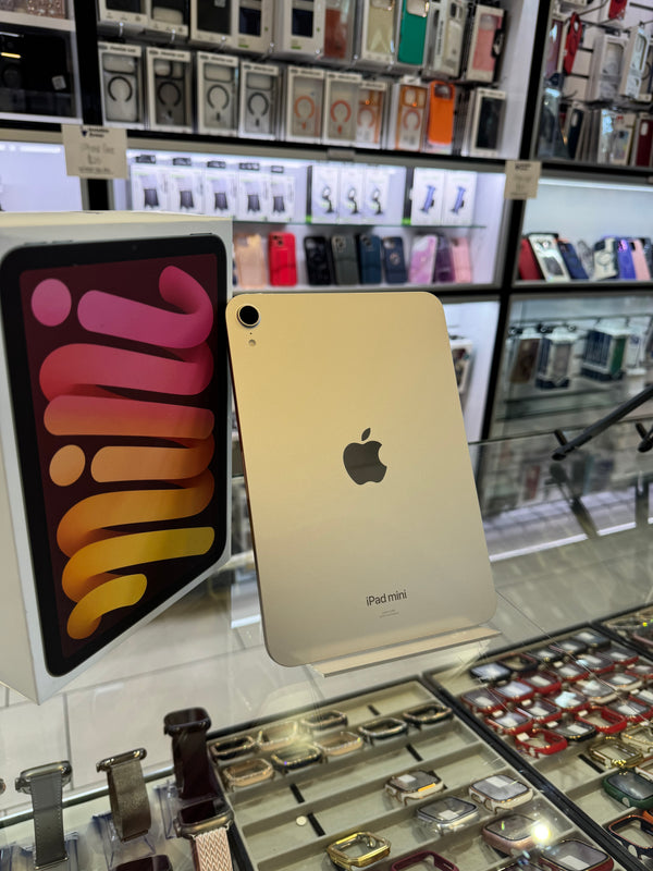 An Apple iPad mini standing upright on a display stand inside a retail store, with its retail box visible to the left and watch and phone accessories in the background.