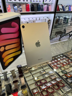 A gold Apple iPad Pro shown standing upright in a retail display, with its retail box partially visible on the left and watch straps and phone cases in the background.
