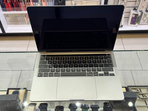 A silver Apple MacBook laptop with a black screen and keyboard is displayed on a glass counter inside a retail store.