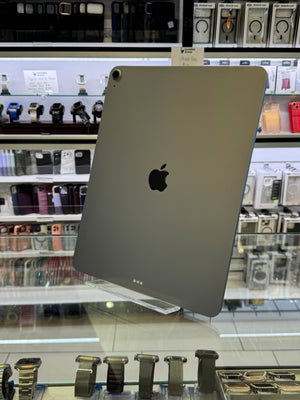 A silver Apple iPad Pro shown from the back, leaning on a clear display stand inside a retail store with shelves of accessories in the background.