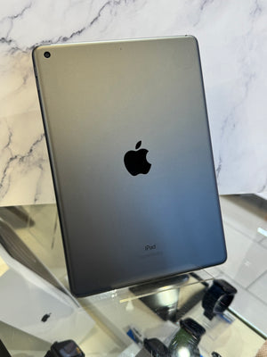 A silver Apple iPad shown from the back, standing on a reflective glass surface against a white marble-patterned background.