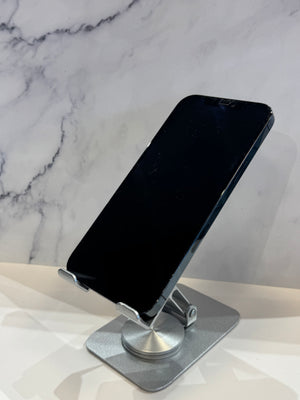 A silver metal adjustable phone stand holding a black smartphone, set against a white marble-patterned background.