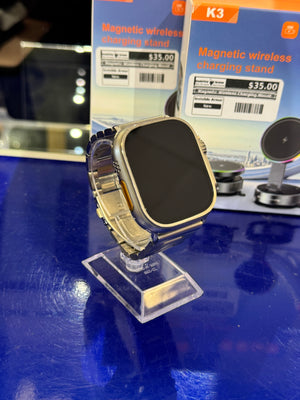 Gold smartwatch on a clear stand with 'K3 Magnetic wireless charging stand' packaging in the background.