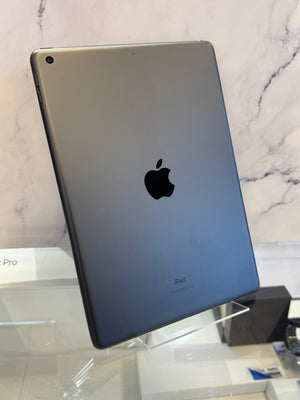 An Apple iPad Pro shown from the back, featuring a dark gray metallic finish with the iconic black Apple logo in the center, displayed on a clear stand against a marble-patterned background.