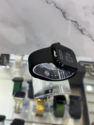 A black smartwatch with a glossy black band, mounted on a clear plastic display stand in a retail setting with marble-patterned background and other watches blurred below.