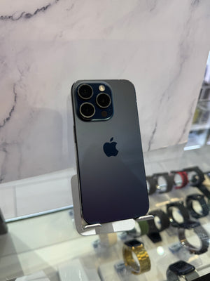 A dark blue-gray Apple iPhone 12 Pro shown from the back, standing on a small display stand in a retail setting with a marble-patterned wall and watch displays in the background.