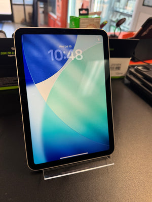 A tablet device with a black bezel and silver edge, standing upright on a clear acrylic display stand on a dark tabletop. The screen is on and displays a blue, green, and white abstract wallpaper with the time 10:48.