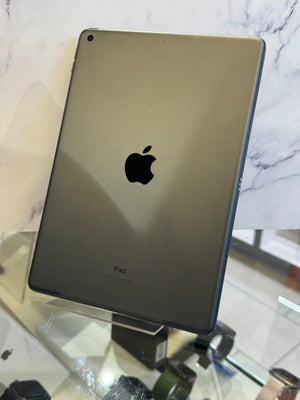 A dark gray Apple iPad shown from the back, resting on a clear display stand on a glass counter with a marble-patterned background.