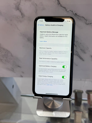 A black smartphone displayed upright on a metal stand, showing the battery health and charging screen. The phone is photographed in a real indoor setting with a marble-patterned wall and blurred objects in the background.