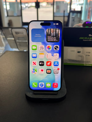 A black smartphone with a lit home screen displaying various app icons, resting on a black charging dock on a dark tabletop. The background shows a blurred retail or office environment with glass windows and display boards.