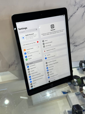 A black tablet device displaying the Settings app on a glass display stand in a retail or showroom environment with marble walls and other devices visible in the background.