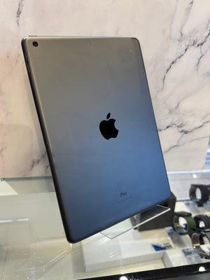 A silver Apple iPad shown from the back, standing on a clear display stand in front of a white marble-patterned wall.