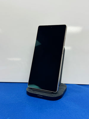 A black smartphone with a silver edge stands vertically on a dark charging dock against a white background with a blue surface below.