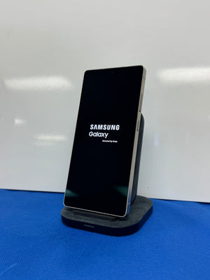 A Samsung Galaxy smartphone with a black screen stands upright on a dark charging dock against a white background, with a blue surface underneath the dock.