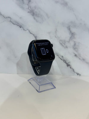 A black Apple Watch with a dark band stands on a clear acrylic display stand against a white marble-patterned background.