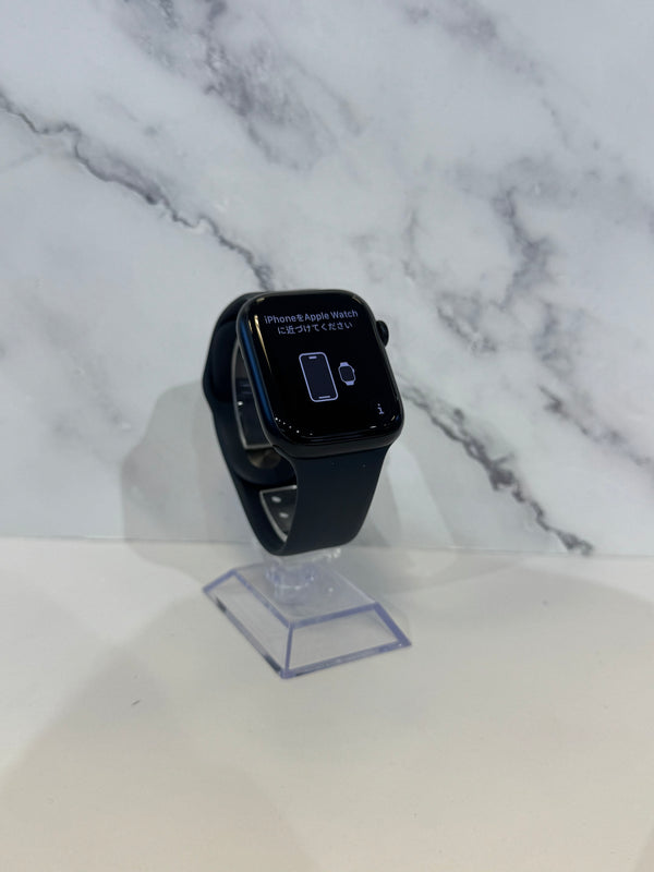 A black Apple Watch with a dark band stands on a clear acrylic display stand against a white marble-patterned background.