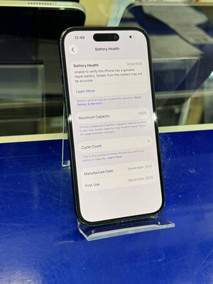 A black smartphone displayed on a clear stand, showing the battery health settings screen on its home screen. The phone is photographed on a blue surface with some reflections and other devices partially visible in the background.