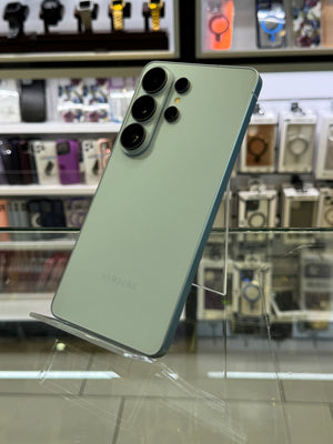 A Samsung smartphone shown from the back, standing on a clear display stand inside a retail store. The phone has a pale mint green finish with a large vertical triple-camera system on the upper left.