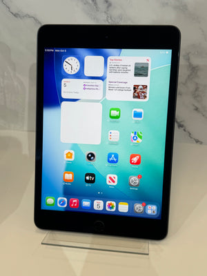 A black Apple iPad tablet standing upright on a clear acrylic display stand, showing its home screen with various apps and widgets. The tablet is photographed on a light beige surface against a white and gray marble-patterned background.