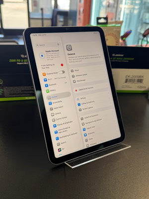 An iPad standing upright on a clear acrylic display stand on a dark tabletop, showing the Settings app screen. The background contains blurred retail packaging and store fixtures.