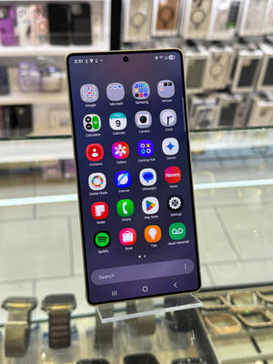 A Samsung smartphone displayed upright on a glass stand inside a retail store, showing its home screen with various app icons. The phone has a black bezel and a dark gray to black front face.