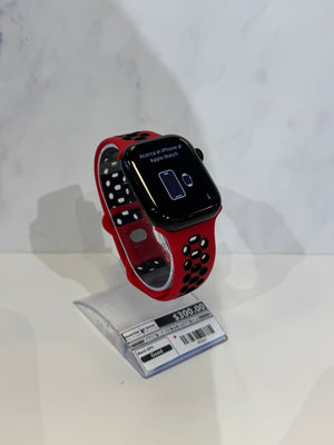A red and black Apple Watch with a red and black perforated sport band, displayed on a clear plastic stand with a price tag in front of it, set against a white marble-patterned background.