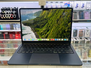 A black Apple MacBook laptop with an open screen displaying a lush green mountain landscape wallpaper, set on a glass display counter with retail accessories in the background.