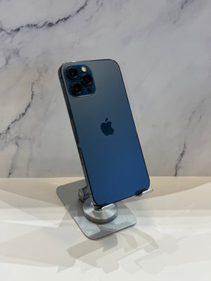 A blue iPhone 12 Pro Max standing upright on a silver metal display stand, showing the back of the phone with its triple-camera array and Apple logo, set against a white marble-patterned background.