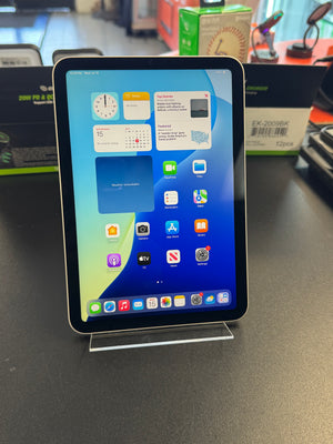 An Apple iPad with a black bezel and silver front face, standing on a clear acrylic display stand on a dark tabletop. The screen is on and displaying the iOS home screen.