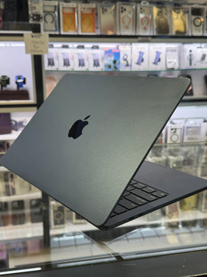 A closed Apple MacBook laptop in dark gray aluminum is shown at an angle on a glass display counter, with a blurred electronics store background.