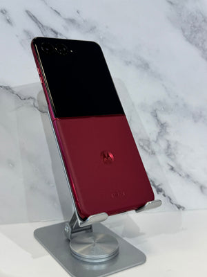 A Google Pixel Fold smartphone with a split black and deep red back panel, displayed on a silver metal stand against a white marble background.