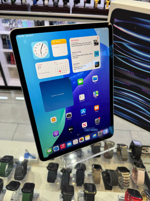 A tablet device with a black bezel and a blue-and-teal home screen, photographed in a retail display area with watches and other electronics in the background.