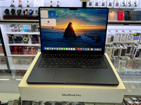 A black Apple MacBook Pro laptop is open and displayed on top of its cream-colored original box, set inside a retail store with shelves of accessories in the background.