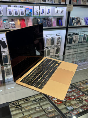 A gold aluminum MacBook laptop with a black screen and keyboard is displayed on a glass counter inside a retail store, with shelves of phone cases and watches visible in the background.