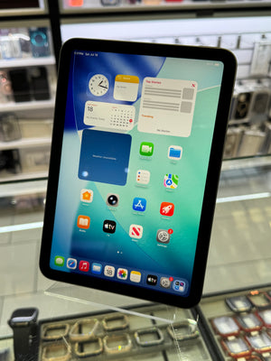 An iPad tablet with a black bezel and colorful home screen, displayed on a clear stand inside a retail store with blurred shelves in the background.
