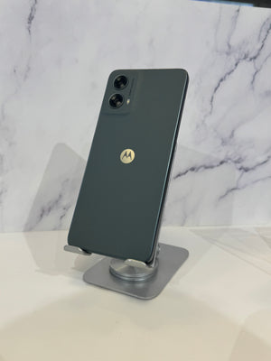 A dark green Motorola smartphone shown from the back, resting on a small metallic display stand against a white marble-patterned background.