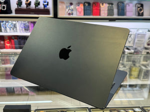 A closed Apple MacBook laptop in dark gray aluminum, shown at an angle on a glass display counter with a blurred electronics store background.