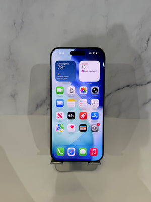 A black iPhone with its screen on, displaying the home screen, resting on a small metal stand against a light gray marble-patterned background.