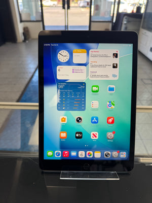 A black Apple iPad tablet standing upright on a clear display stand, showing the home screen with various app icons. The tablet is photographed indoors on a dark counter with a blurred store or office background.